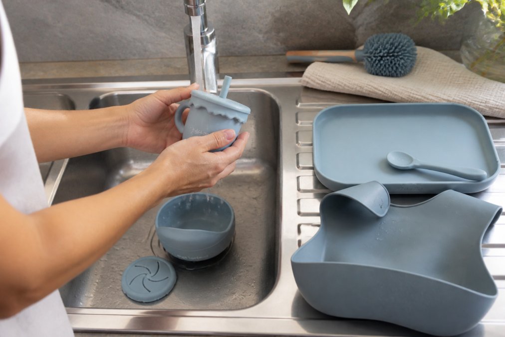 Hands rinsing silicone feeding pieces under warm water in a kitchen sink, soft brush nearby, silicone items appearing vibrant and stain-free, demonstrating easy cleaning process and material durability