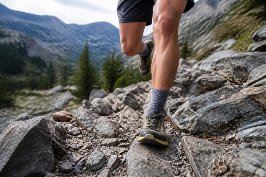 Trail runner on a technical rocky descent, feet visible in protective athletic socks, demonstrating proper footwear for challenging terrain with sharp rocks visible below