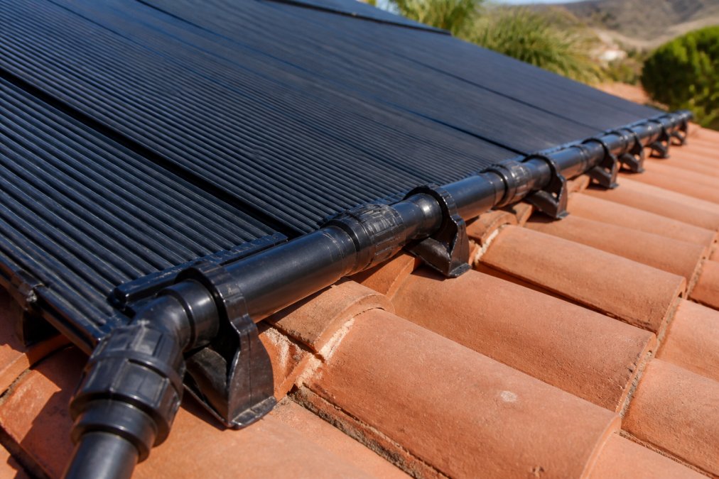 Close-up of polypropylene solar pool heating panels mounted on a residential rooftop, showing the dark panel surface texture and snap-fit connection fittings under bright afternoon sunlight, with terracotta roof tiles visible beneath