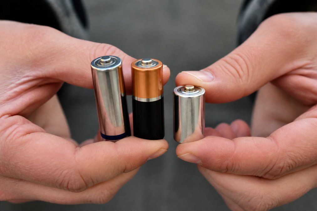 Close-up of hands holding different battery types side-by-side, displaying AA lithium, AA alkaline, and CR123A lithium batteries against a neutral background to show size and form factor differences