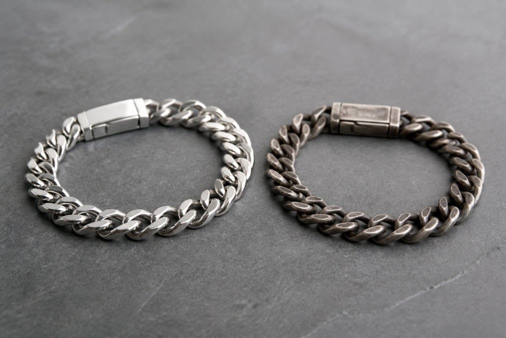 Two pieces of jewelry side by side on a neutral surface, a polished stainless steel bracelet and a tarnished sterling silver bracelet, highlighting their different finishes and color differences after extended wear
