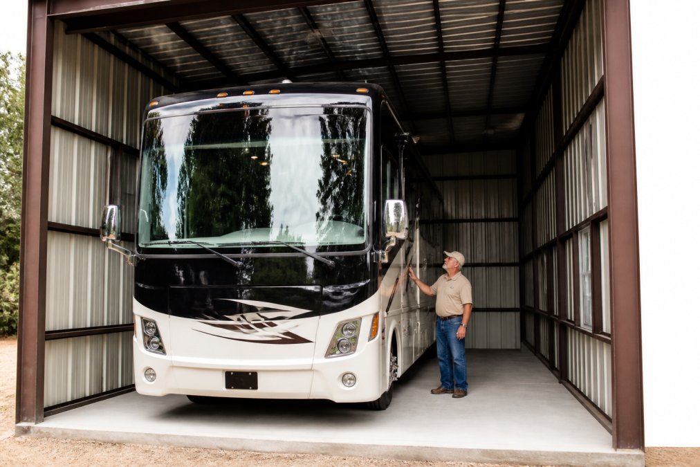A large Class A motorhome parked inside a tall, fully enclosed steel garage, with wide side-wall height and a person standing next to the driver's door for scale