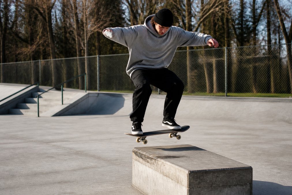 Skateboarder in mid-trick wearing oversized grey hoodie and black joggers, demonstrating relaxed fit and unrestricted arm movement on concrete skate spot with natural daylight