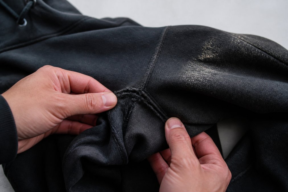 Hands carefully inspecting seams and fabric of worn black hoodie, showing griptape wear patterns and areas needing repair against neutral background