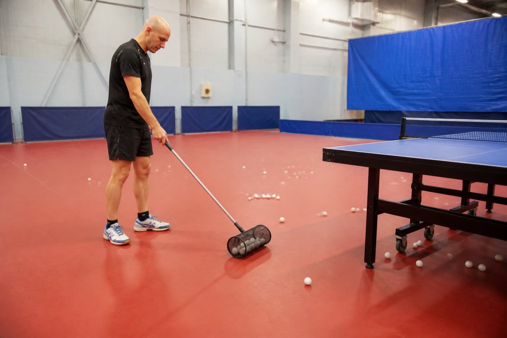 A coach in a bright training hall using the MyPingPongBuddy ball collector to scoop up table tennis balls from the floor without bending over, natural overhead lighting