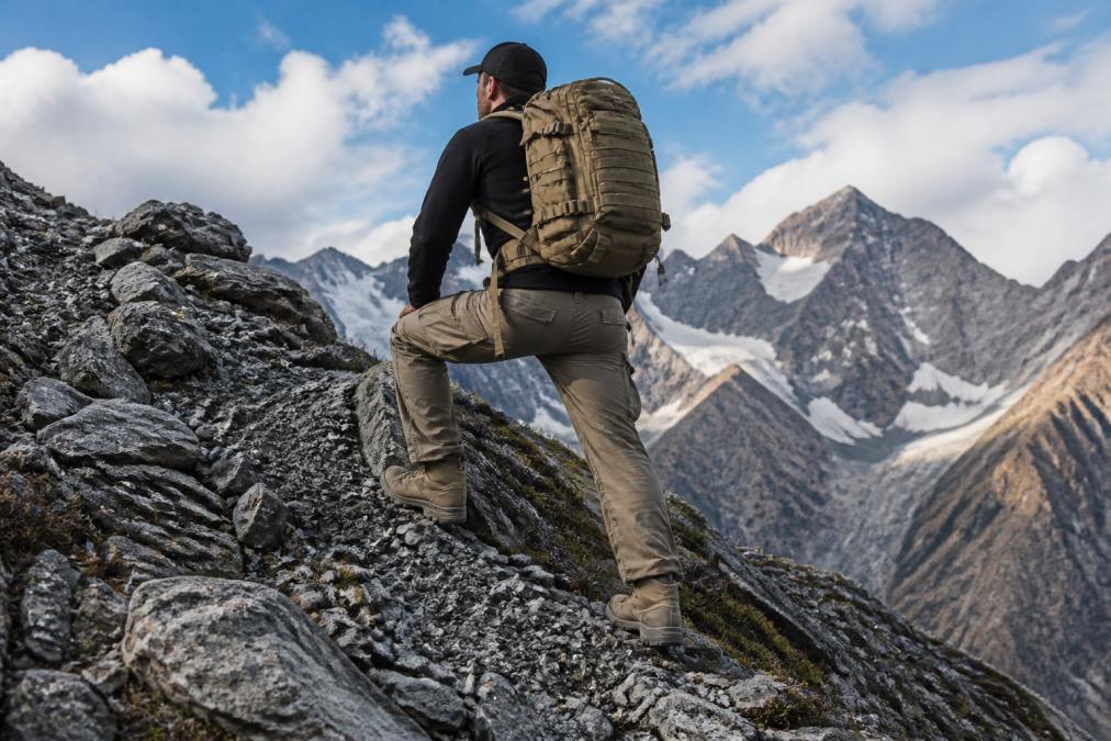 Hiker wearing a tactical backpack and reinforced ripstop pants navigating rocky alpine terrain with steep mountain backdrop, demonstrating gear durability in challenging conditions