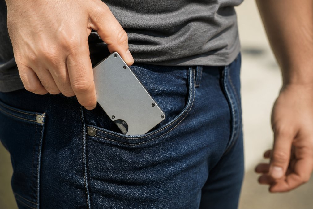 Close-up of hands sliding a slim aluminum tactical wallet out of a jeans pocket, bright daylight showing the brushed metal finish