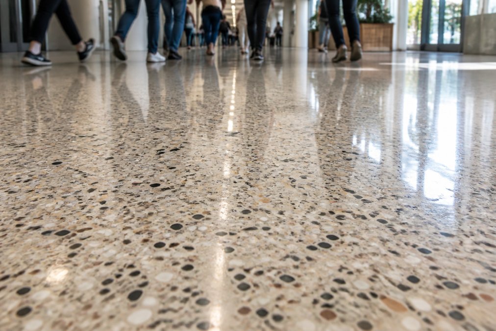 Close-up of a polished terrazzo floor surface in a busy commercial hallway with foot traffic visible, showing the aggregate pattern and reflective finish under bright overhead lighting