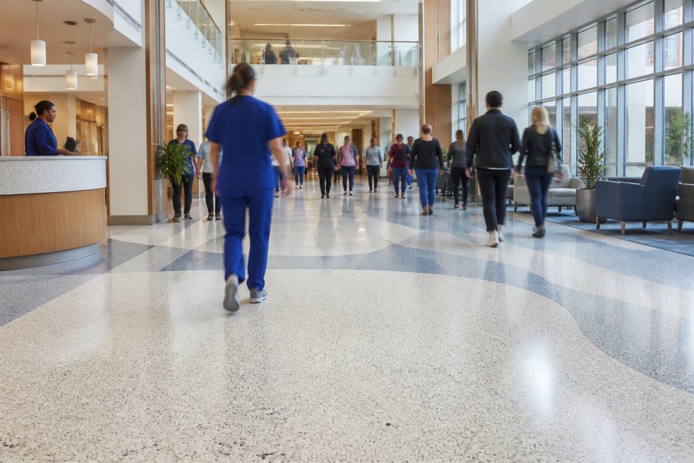 Wide-angle view of a modern hospital corridor or airport terminal with terrazzo flooring, showing crowds of people moving through the space and the floor's durability under heavy use with bright, clean lighting