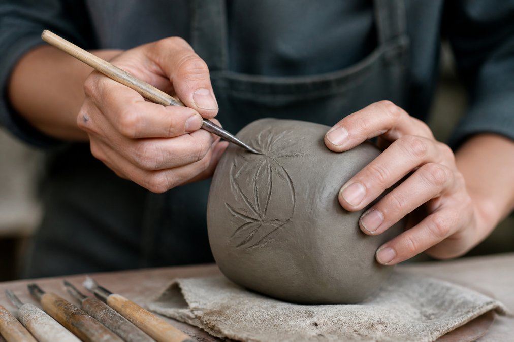 Close-up of ceramic artist's hands using carving tools on leather-hard clay, showing proper grip and detailed incising work with natural studio lighting