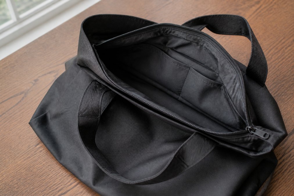 A large polyester tote bag laid flat on a wooden table in natural daylight, showing open interior pockets, a visible zipper closure, and reinforced stitching at the handles