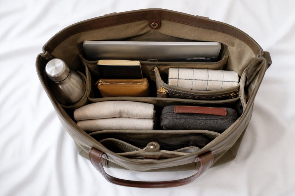Overhead flat-lay of a tote bag open on a white surface, contents neatly arranged with a laptop visible in the back sleeve, a water bottle in the side pocket, and a passport and small pouch grouped near the top opening, clean minimalist styling in natural light