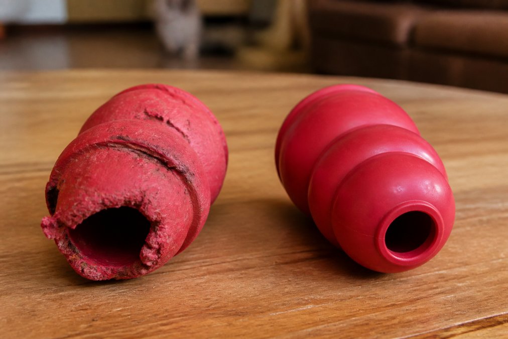 Close-up of a visibly worn and cracked rubber chew toy with missing chunks placed next to an intact replacement toy on a wooden surface, under warm indoor lighting