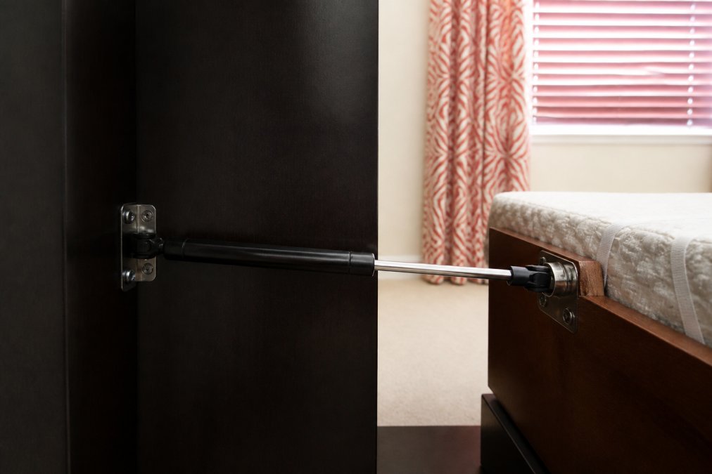 Close-up of a gas-strut piston cylinder mounted horizontally on wall bed frame hardware, showing the metal rod and articulating connection point in a modern bedroom setting