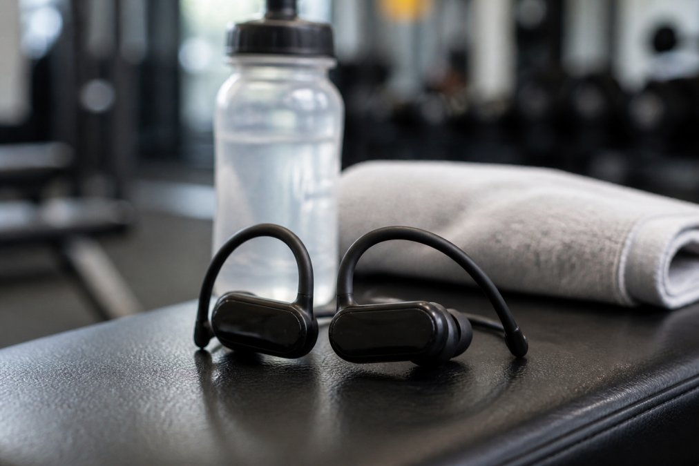 Close-up of black Bluetooth 5.3 ear hook earbuds resting on a gym bench with a water bottle and towel blurred in the background