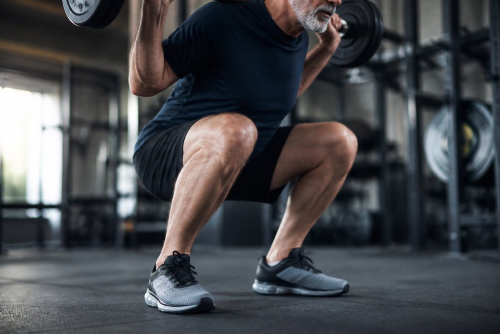 Older athlete's feet in supportive workout shoes during a weighted squat movement, showing proper foot positioning and arch support with visible cushioning under the heel and forefoot