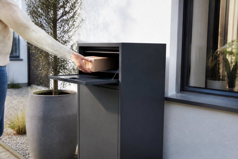 Een hand plaatst een klein pakketje in de geopende klep van een donkergrijze, vrijstaande pakketbrievenbus voor een moderne witte gevel, met zacht ochtendlicht dat over het gepoedercoate staal valt