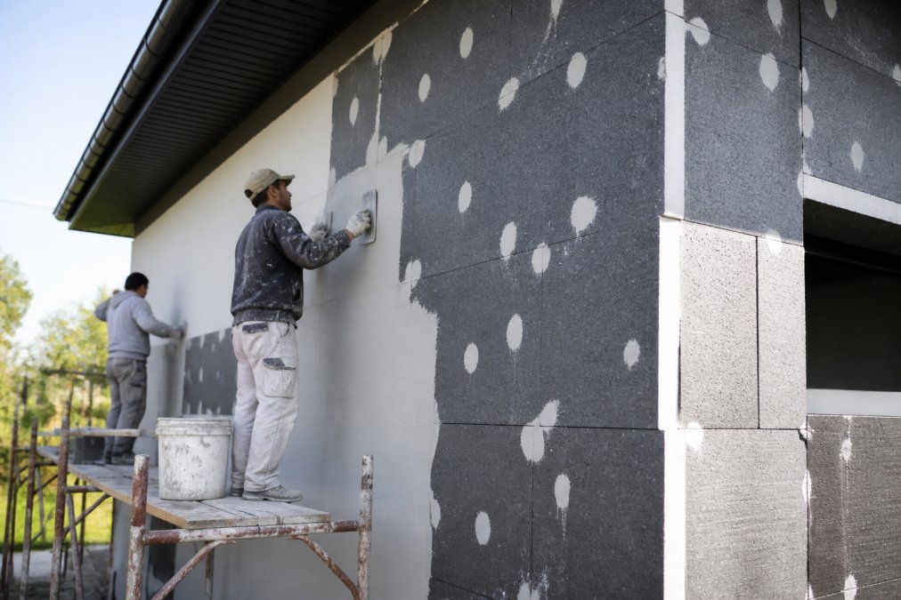 Professionele werkers voeren gevelisolatie uit op een woning, zichtbaar isolatieplaten tegen de muur bevestigd, met verflaag en afwerkingsmateriaal, daglicht op de werkplek