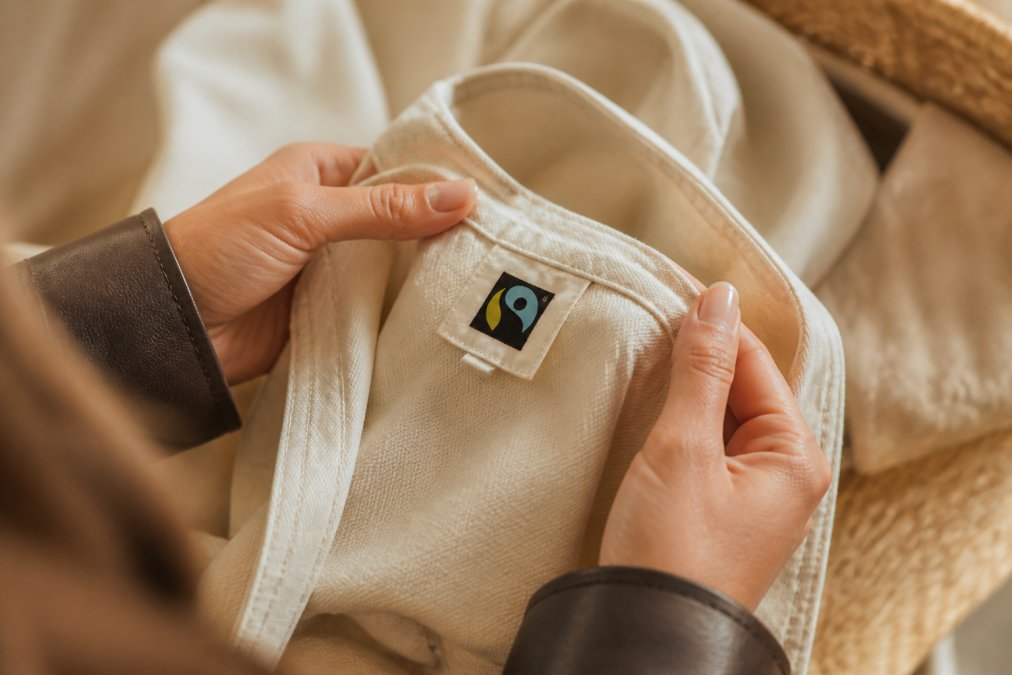 Close-up van handen die een natuurlijk katoenen kledingstuk voorzichtig controleren met een Fair Trade keurmerk duidelijk zichtbaar op het label, tegen een zachte beige achtergrond met natuurlijk daglicht