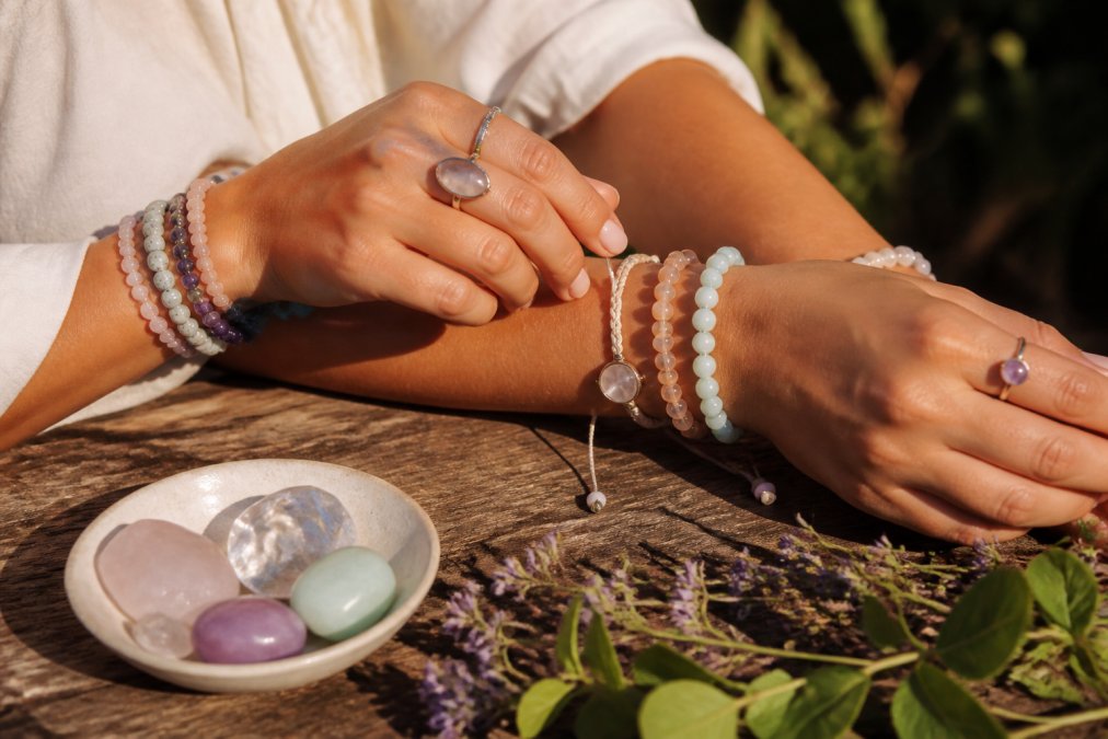 Close-up van iemands polsen en handen met verschillende spirituele armbanden en ringen met natuurlijke edelstenen in zacht zonlicht, rustende op een houten tafel in een serene natuuromgeving
