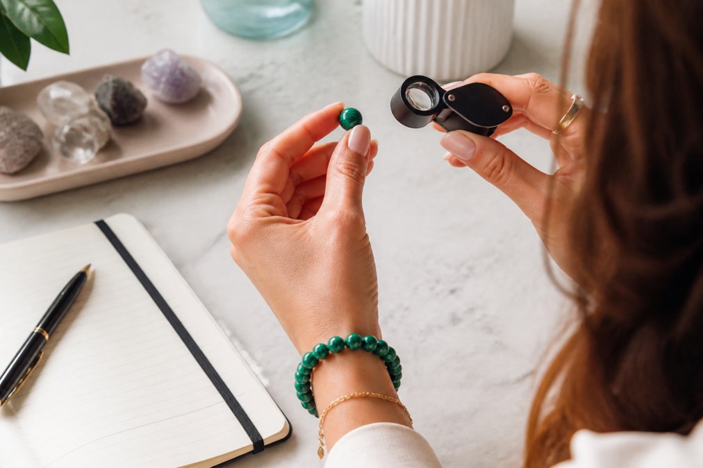 Handen van een vrouw die een losse edelsteen vasthoudt en nauwkeurig bekijkt, met een vergrotingsglas en een klein notitieboekje op een lichte marmeren tafel