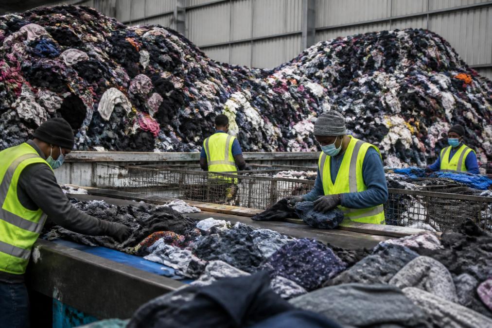 Textielafval en oude kleding gestapeld in grote hoeveelheden op een recyclingfaciliteit, met werknemers in reflecterende vesten die sorteren en controleren