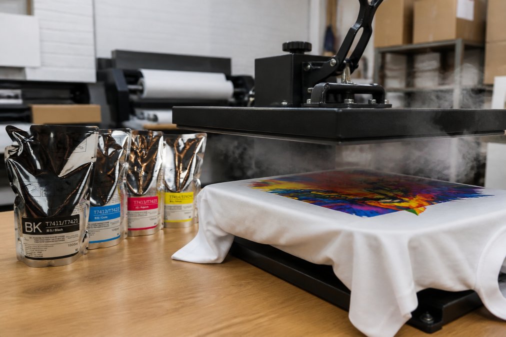 Close-up of a heat press machine pressing a vibrant full-color design onto a white polyester shirt on a worktable in a print shop, with steam rising from the press under bright workshop lighting