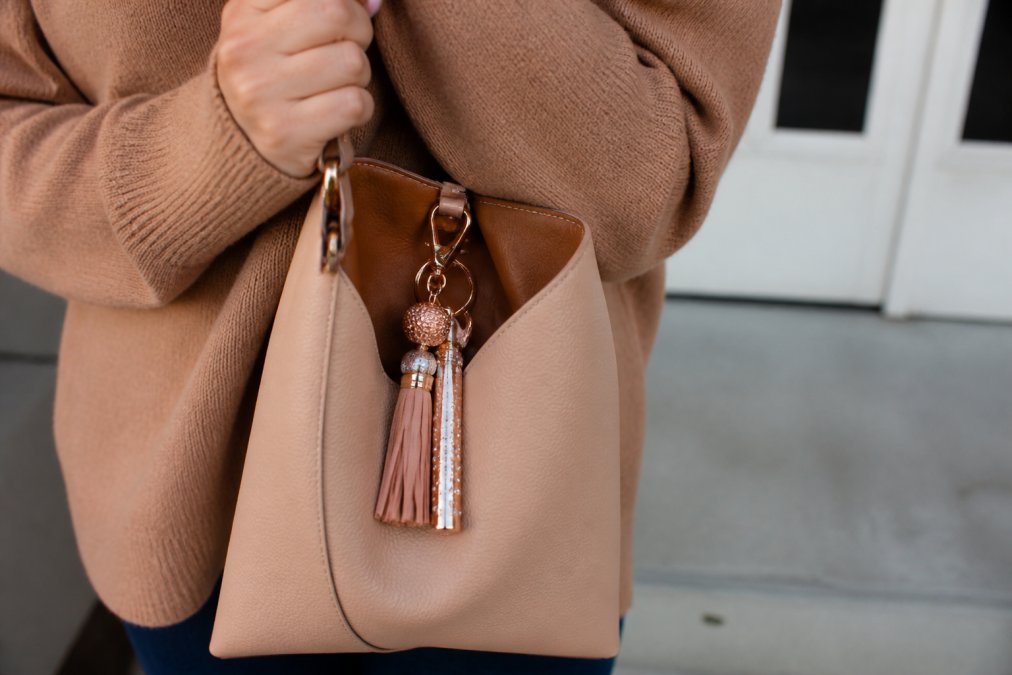 Close-up of a woman's hand holding a designer handbag with a stylish keychain accessory clipped to the interior key ring, showing coordinated warm metallics and complementary leather tones in natural daylight