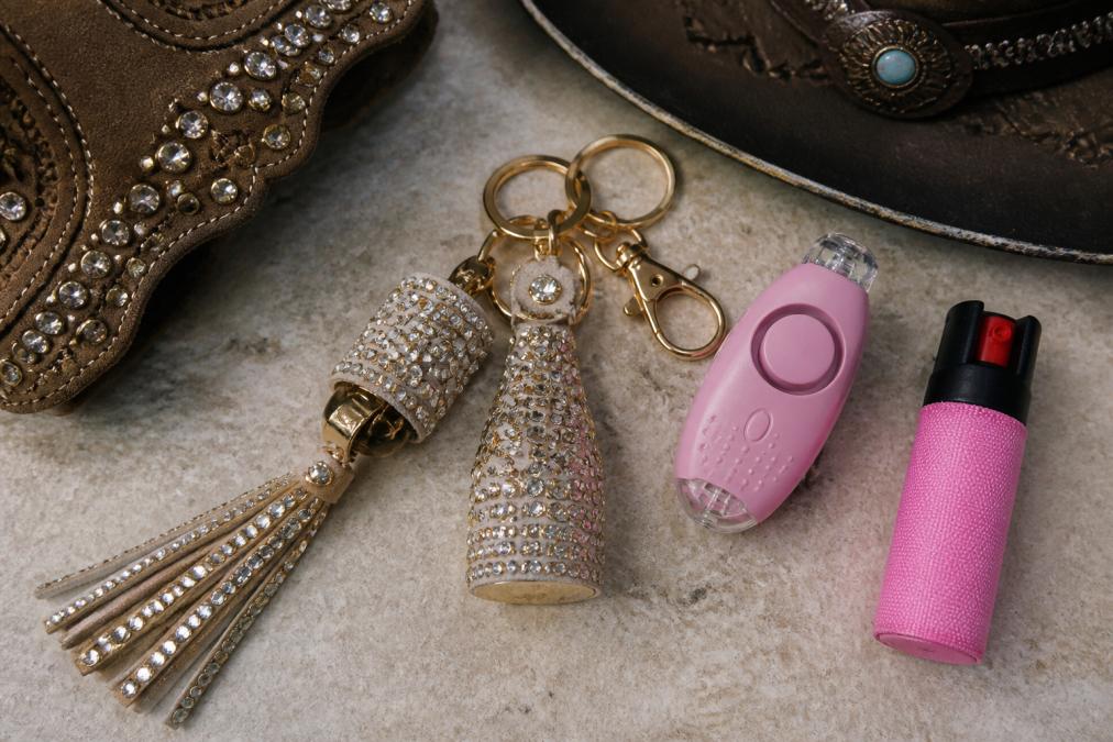 Flat-lay arrangement showing a designer rhinestone safety keychain with matching Western-inspired accessories, personal alarm with LED light, and compact pepper spray arranged on neutral background with soft natural lighting