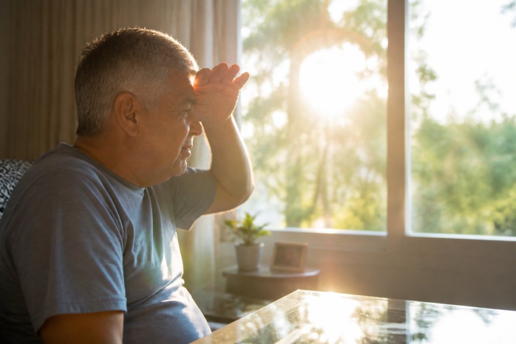 Mature adult sitting in a bright home environment, squinting toward a window with visible glare and light sources creating a halo effect