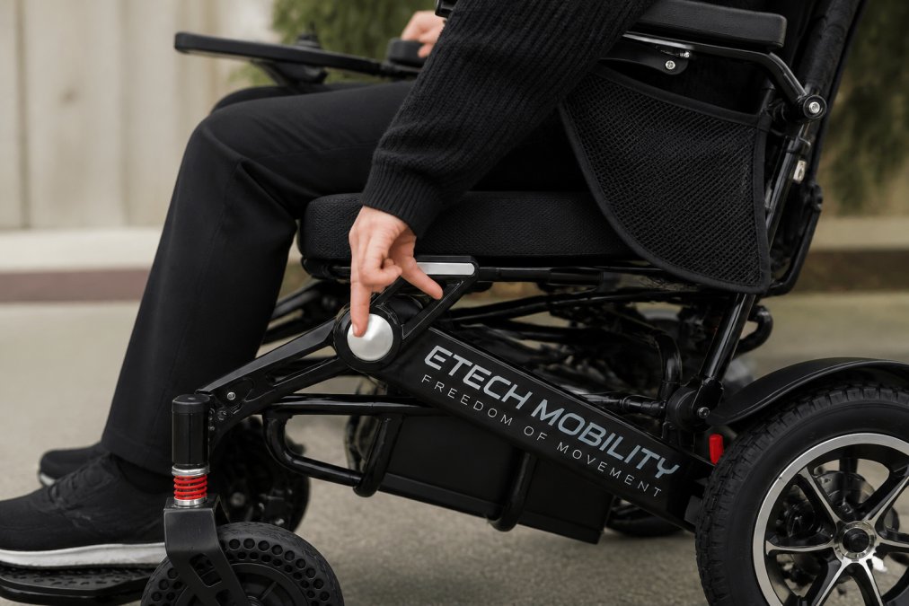 Close-up of hands gently operating an automatic fold button on a lightweight electric wheelchair, showing comfortable hand positioning without strain