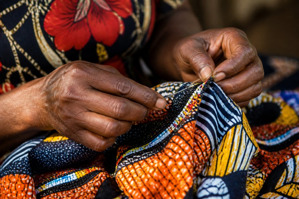 Close-up of skilled artisan hands carefully hand-stitching vibrant African print fabric with traditional needle work, showing detailed beadwork and intricate stitching patterns under warm natural light