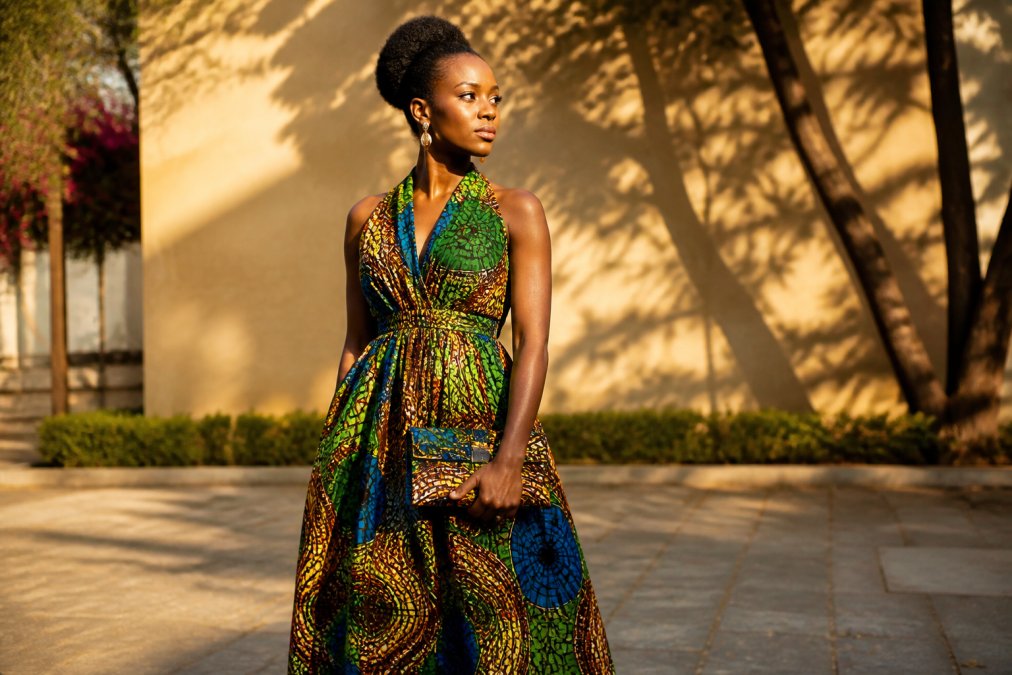Woman wearing a vibrant handcrafted African print infinity dress with matching structured clutch, photographed outdoors in warm natural sunlight showcasing the rich colors, intricate patterns, and quality construction of the garment