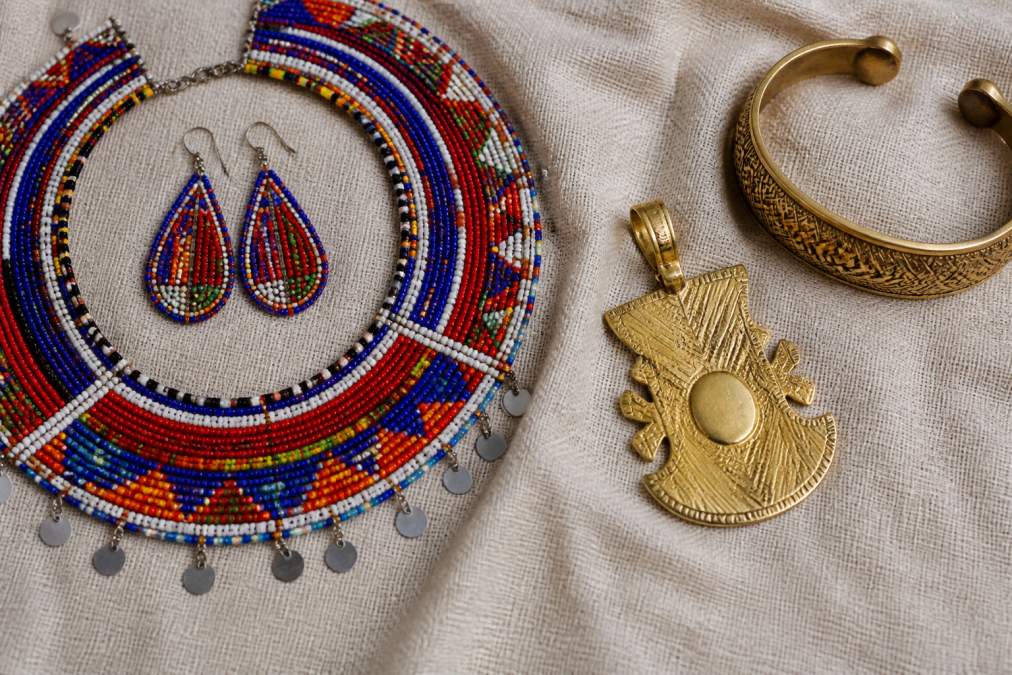A flat lay of two distinct jewelry styles on neutral linen fabric: on the left, a vibrant Maasai beaded collar and seed bead earrings in red, blue, and green; on the right, a polished gold pendant and brass bracelet with intricate carved patterns