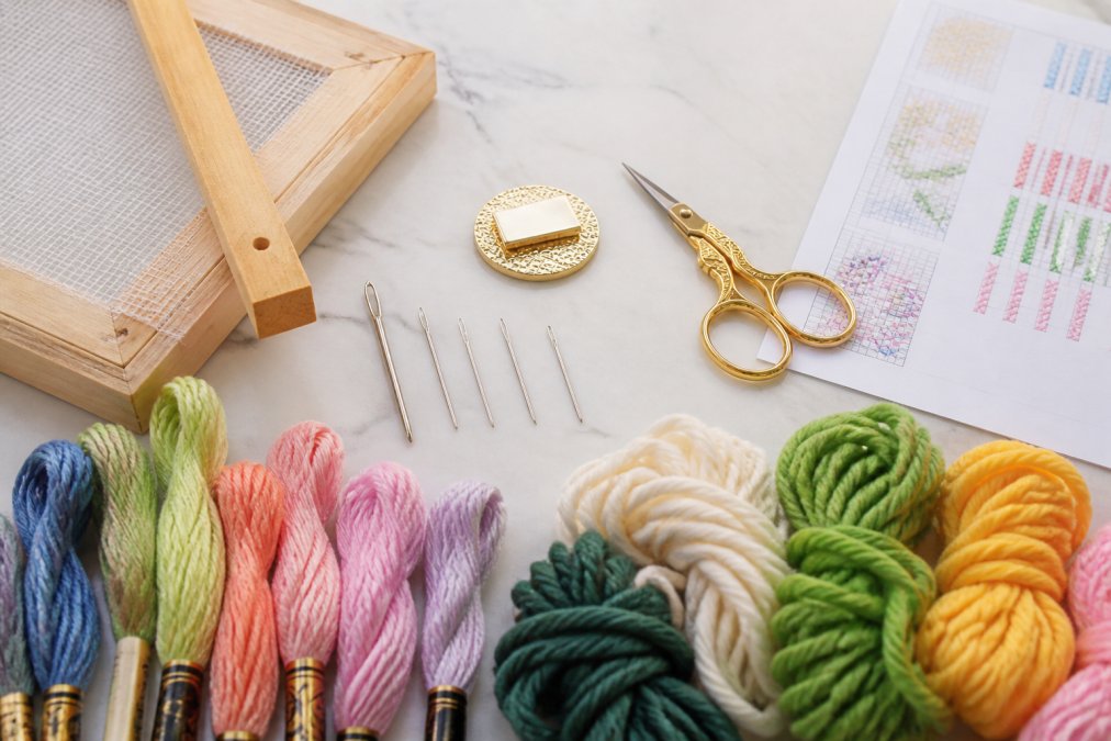 Flat lay arrangement of needlepoint supplies on light background: tapestry needles in various sizes, embroidery floss and tapestry wool in multiple colors, stretcher bars with canvas attached, needle minder, embroidery scissors, and stitch diagram reference sheet