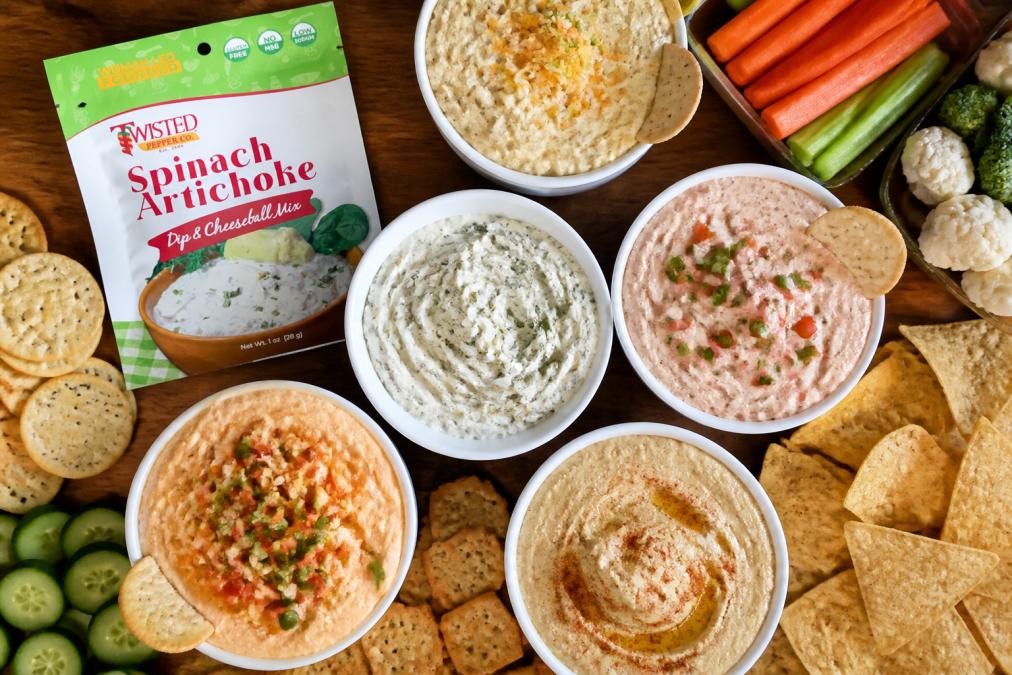 Overhead shot of five colorful dips in white bowls arranged on a wooden table with tortilla chips, crackers, and fresh vegetables scattered around for dipping