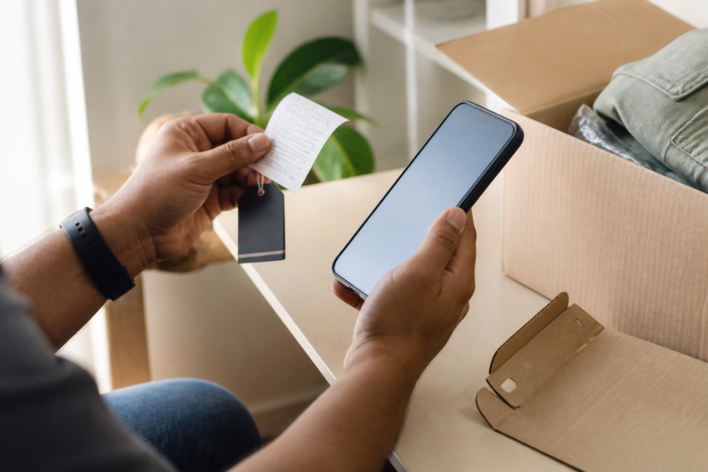Close-up of hands holding a product receipt and original tags while reviewing return policy details on a smartphone screen, with a package and original packaging visible in soft natural light
