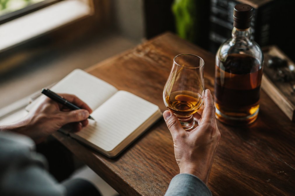 A person's hands holding a whiskey glass with a notebook and pen nearby, capturing tasting notes with a bottle visible in the background