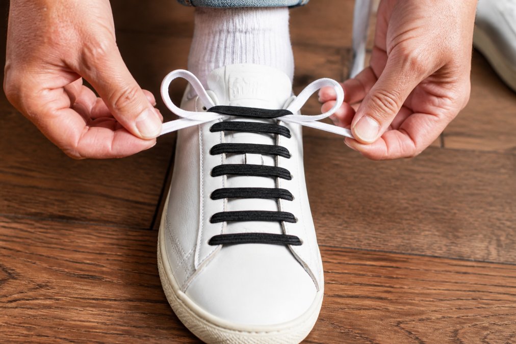 Close-up of an adult's hands forming two loops with white shoelaces on a classic sneaker, demonstrating the bunny ears tying method on a wooden floor