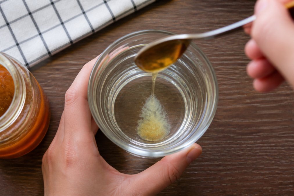 Hände führen einen Wasserlöslichkeitstest mit Honig durch - Glas mit klarem Wasser und Honig wird von oben fotografiert, der Honig sinkt langsam zu Boden und bildet eine milchige Trübung, natürliches Licht