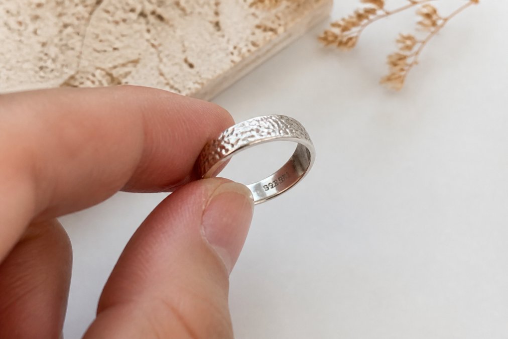 Close-up of a silver ring held between two fingers, showing a tiny 925 hallmark stamp engraved on the inner band, soft natural lighting on a white surface