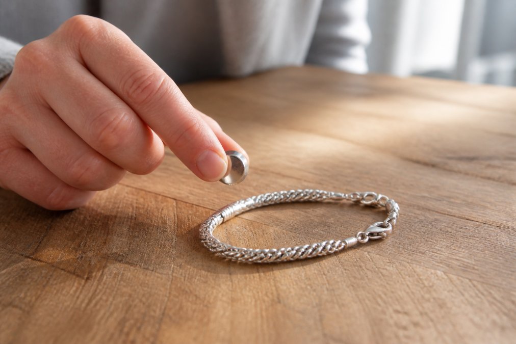 A hand holding a small neodymium magnet near a silver bracelet resting on a wooden table, demonstrating the magnet test for silver authenticity, warm natural window light