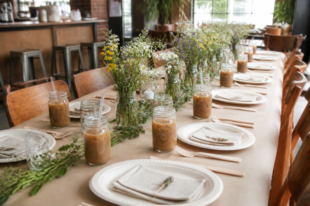 A beautifully set bridal shower table in a bright cafe with mason jar centerpieces holding wildflowers, compostable dinnerware, kraft paper runners, and glass iced coffee cups at each place setting