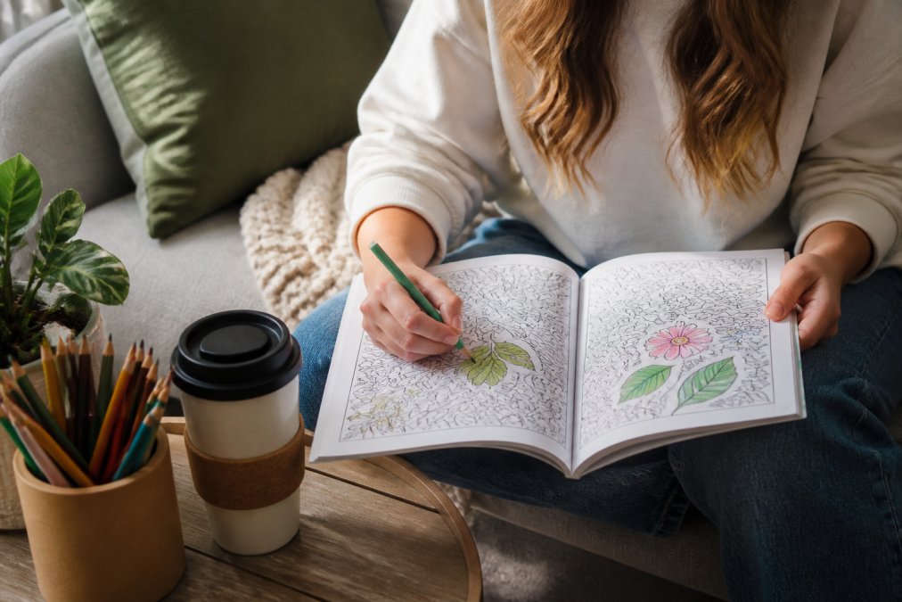 A person relaxing on a couch, using colored pencils on an open adult coloring book with botanical designs, with a reusable coffee mug nearby