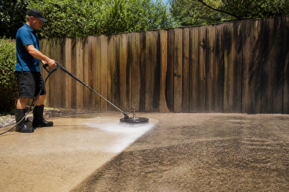 Professional demonstrating eco-friendly exterior cleaning technique on concrete driveway using low-pressure equipment with visible water spray, timber fence in background