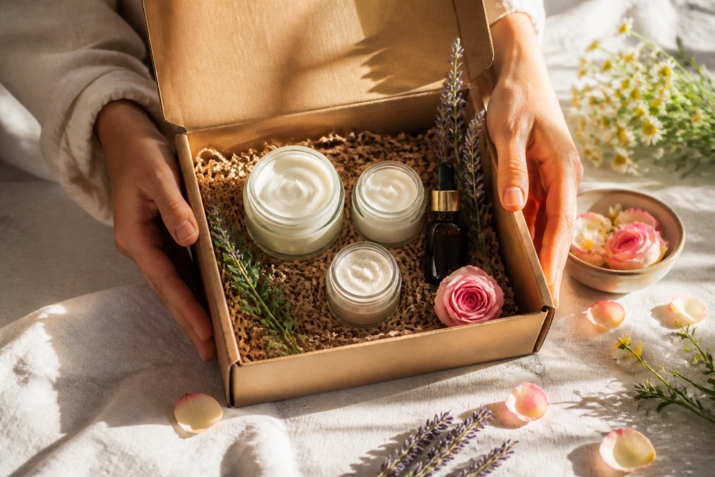Hands gently opening a natural gift box containing glass jars of organic skincare products surrounded by fresh botanical elements like lavender sprigs and rose petals, with soft morning sunlight streaming across the scene