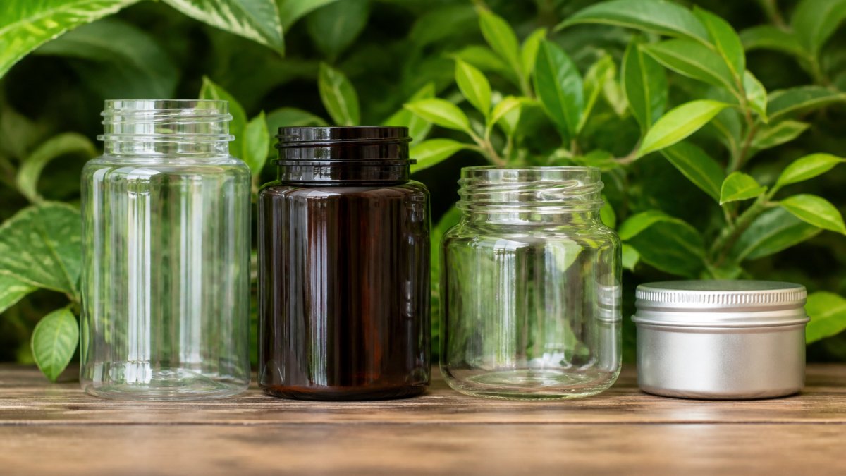 Clear plastic, amber HDPE, glass, and aluminum containers arranged with green leaves on a wooden surface.