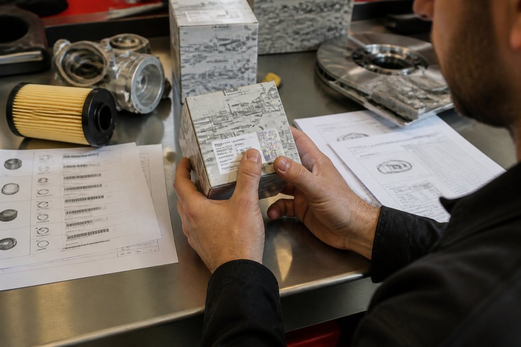 Hands of a technician carefully examining OEM part number labeling and hologram authentication on genuine Audi component packaging, with technical specification sheets and compatibility documentation visible on a clean workbench
