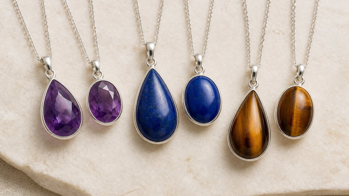 Nahaufnahme mehrerer Edelstein-Anhänger in tropfenförmiger und ovaler Form mit unterschiedlichen Farben (Amethyst violett, Lapislazuli blau, Tigerauge braun) an feiner Silberkette, auf neutralem cremefarbenem Hintergrund