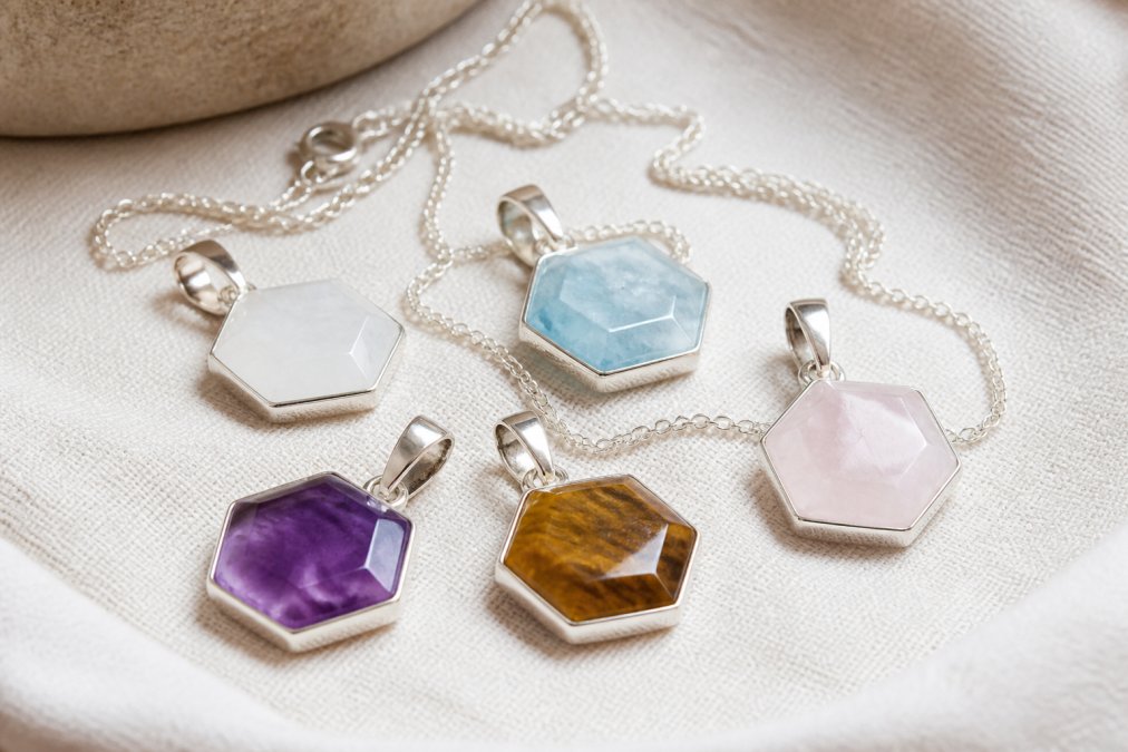Bunte, sechseckige Edelstein-Anhänger und eine Silberkette als edelsteinschmuck online auf hellem Leinenstoff präsentiert.